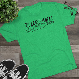 Green tri-blend "Tiller Mafia" graphic tee with black sneakers, plant, and glasses on wood.