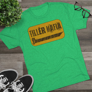 Green "TILLER MAFIA" tee with yellow and black graphic, black sneakers, plant, and glasses on wood.