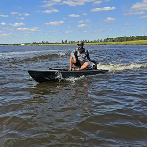 Man in a camo shirt on an olive Crescent Cres-Craft Power Kayak with a motor, speeding on water.