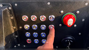 Hand presses a 22mm Plashlights REVERT marine switch with a red LED ring on a black panel.