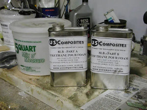 Two silver US Composites 8lb urethane pour foam cans (Part A & B) next to a white 5-quart measuring bucket.