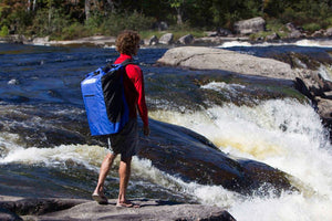 Man wearing a large blue and black Sea Eagle backpack with padded straps, standing by a river with rapids.