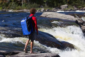 Man wearing a large blue and black Sea Eagle backpack with padded straps, standing by a river with rapids.