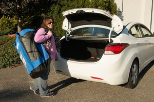 Woman carrying a large blue Sea Eagle backpack with mesh panels towards an open white car trunk.
