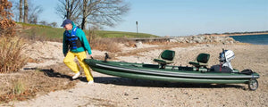 Man in blue vest and yellow pants carrying a green Sea Eagle FishSkiff 16 inflatable fishing boat with two seats and an outboard motor on a sandy beach.