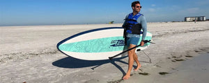 Man in a life vest carrying a white, blue, and teal Sea Eagle LongBoard 11 inflatable paddleboard and paddle on a sandy beach.