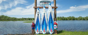 Three blue and white Sea Eagle NeedleNose™ inflatable paddleboards with paddles lean against a wooden marina sign.