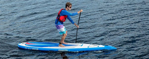 Man paddling a blue and white Sea Eagle NeedleNose 126 inflatable paddleboard on calm water.