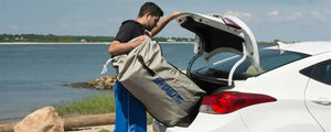 Man loading a compact, rolled-up Sea Eagle inflatable boat in a gray storage bag into a white car trunk by the water.