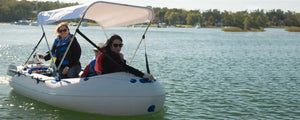 Three people in a light gray Sea Eagle 9 inflatable boat with a white canopy on calm water.
