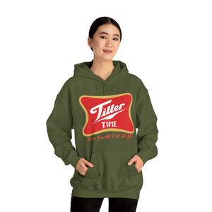 Woman wearing an olive green "Tiller Time" graphic hoodie with a red and gold emblem, hands in kangaroo pocket.