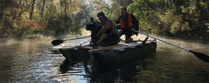 Two men in a dark hunter green Sea Eagle 375fc FoldCat inflatable pontoon fishing boat with oars on a misty river.