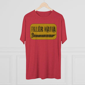 A red short-sleeve t-shirt hanging on a light wood hanger against a white paneled wall. The shirt features a yellow graphic on the chest with the text 'TILLÉR MAFIA' in black.