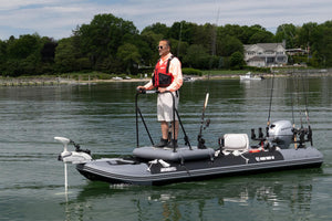 A man stands on a gray sea eagle inflatable fishing boat with a mounted motor, fishing rods, and a white outboard motor, on the water with a shoreline, trees, and houses in the background.