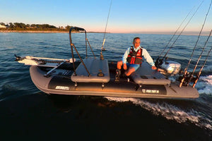 A man wearing a life jacket sits on a gray sea eagle inflatable fishing boat with multiple fishing rods mounted on the sides, moving across open water during sunset.