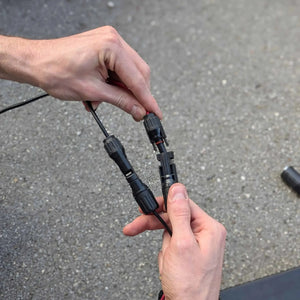 Two hands holding black electrical connectors against a gray pavement background