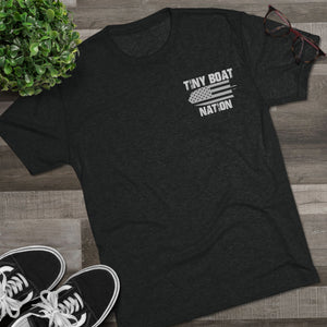 Black t-shirt with 'Tiny Boat Nation' design on a wooden surface with sneakers and a plant.