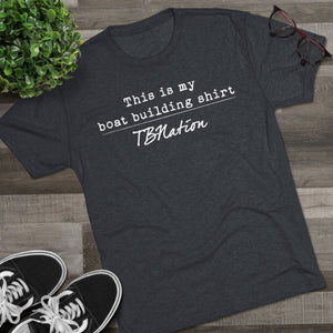 Dark gray t-shirt with text on a wooden surface with sneakers and a plant.
