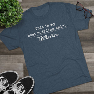 Blue t-shirt with text on a wooden surface with shoes and a plant.
