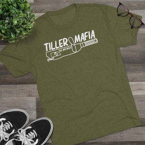 An olive green heather t-shirt with a white graphic reading 'TILLER MAFIA' and 'JON YACHT' over a baseball bat illustration, styled in a flat lay on a wood floor with black sneakers, glasses, and a green plant.