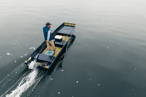 Man in a small, black flat-bottom boat with a white outboard motor, moving across calm water.