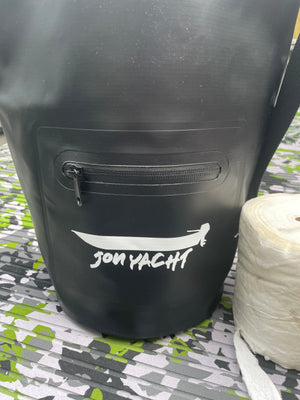 Black 5L Jon Yacht dry bag with a zippered front pocket and white logo, next to a roll of white material.
