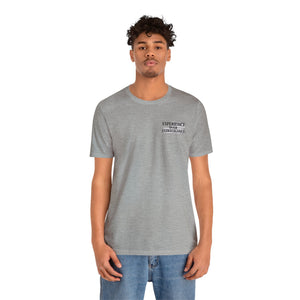 Man wearing a light gray "American Dreamer" unisex jersey tee with "EXPERIENCE OVER EXTRAVAGANCE" text.
