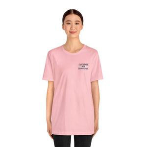 Woman wearing a light pink "American Dreamer" unisex jersey short sleeve tee with "EXPERIENCE OVER EXTRAVAGANCE" text.