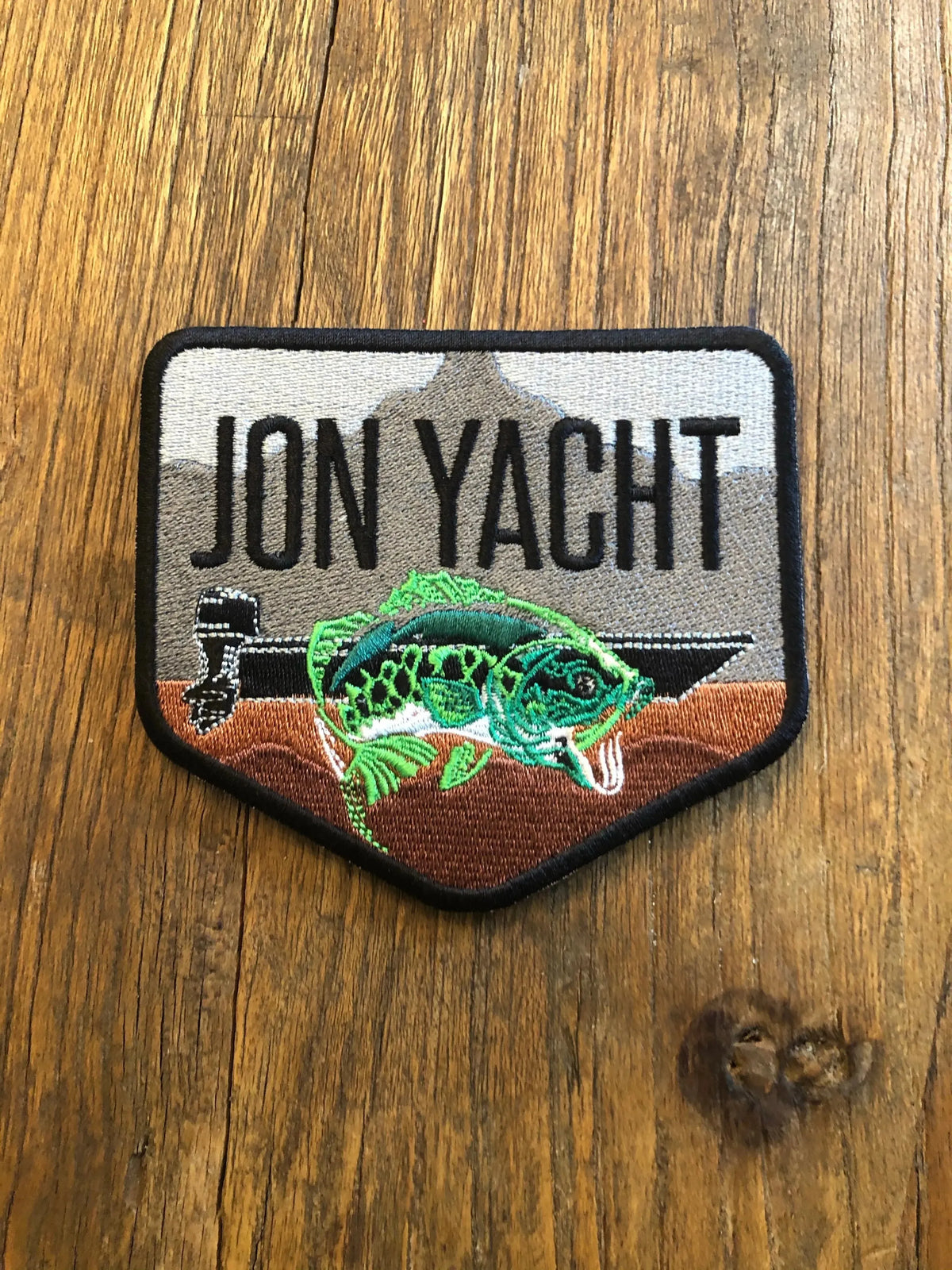 Embroidered Jon Yacht bass patch with a green fish, boat, and mountains on a brown wood background.