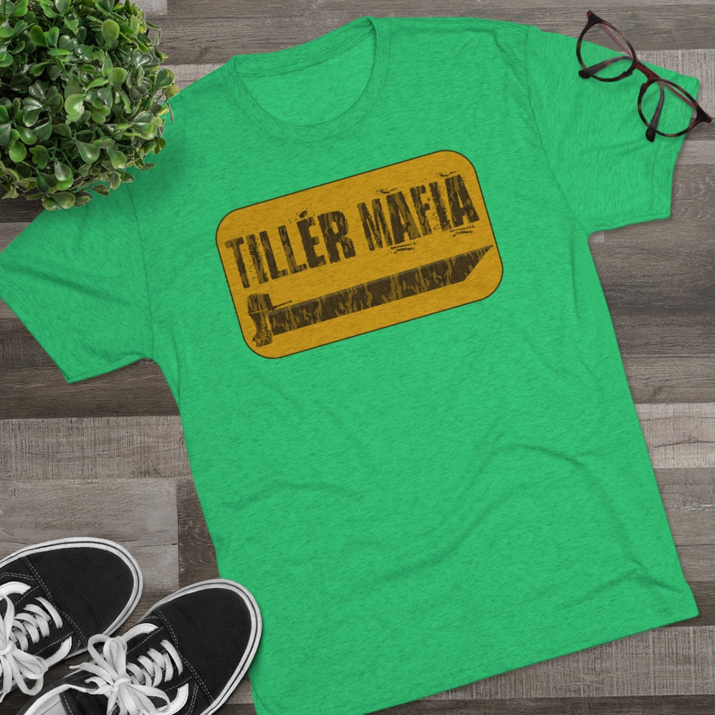 Green "TILLER MAFIA" tee with yellow and black graphic, black sneakers, plant, and glasses on wood.