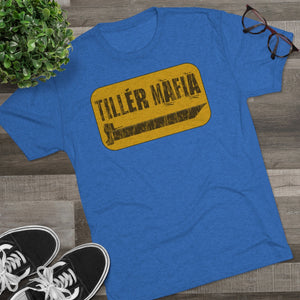 Blue CAMO Tiller Mafia Tee with distressed yellow graphic and tiller blade design, shown on a wooden surface.