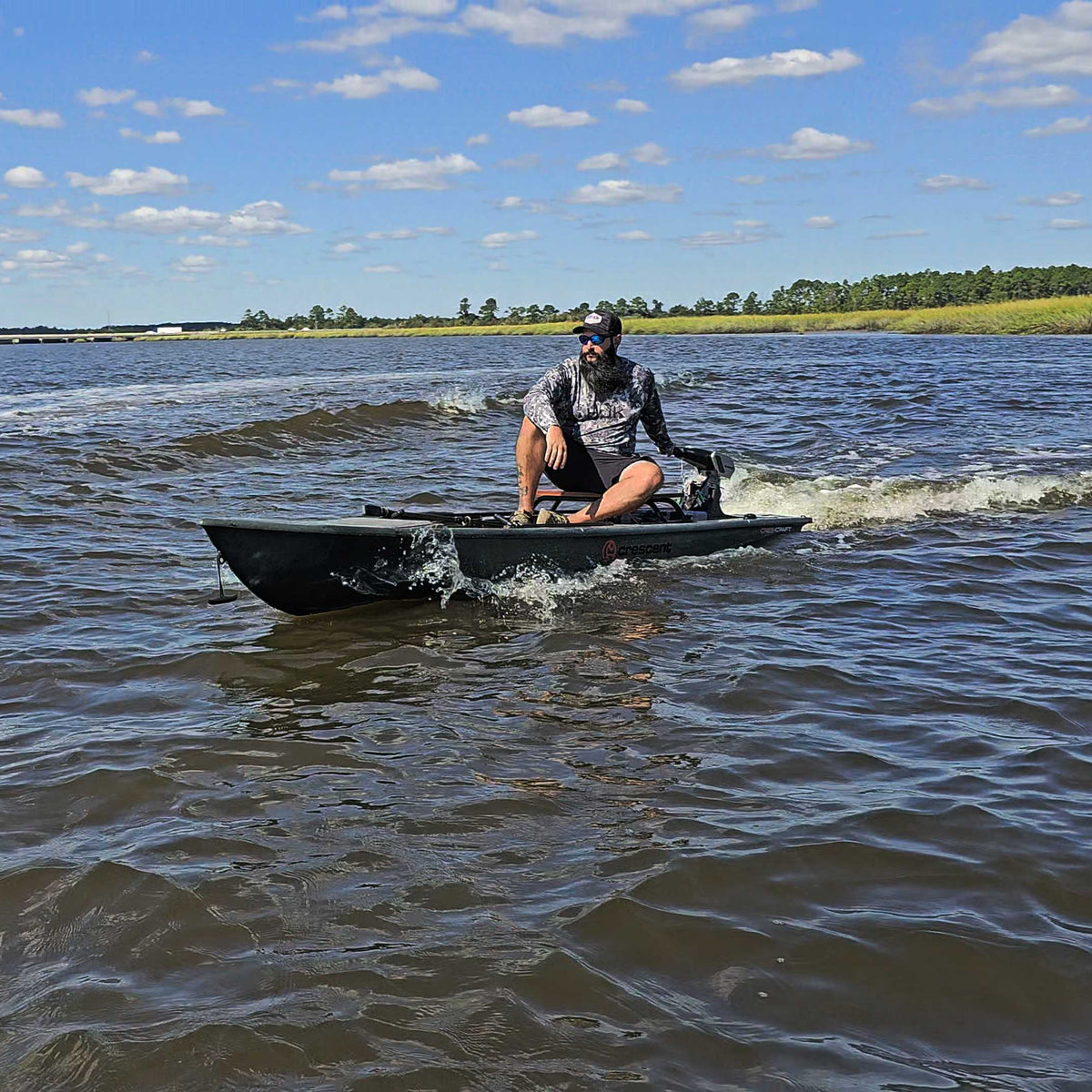 Man in a camo shirt on an olive Crescent Cres-Craft Power Kayak with a motor, speeding on water.