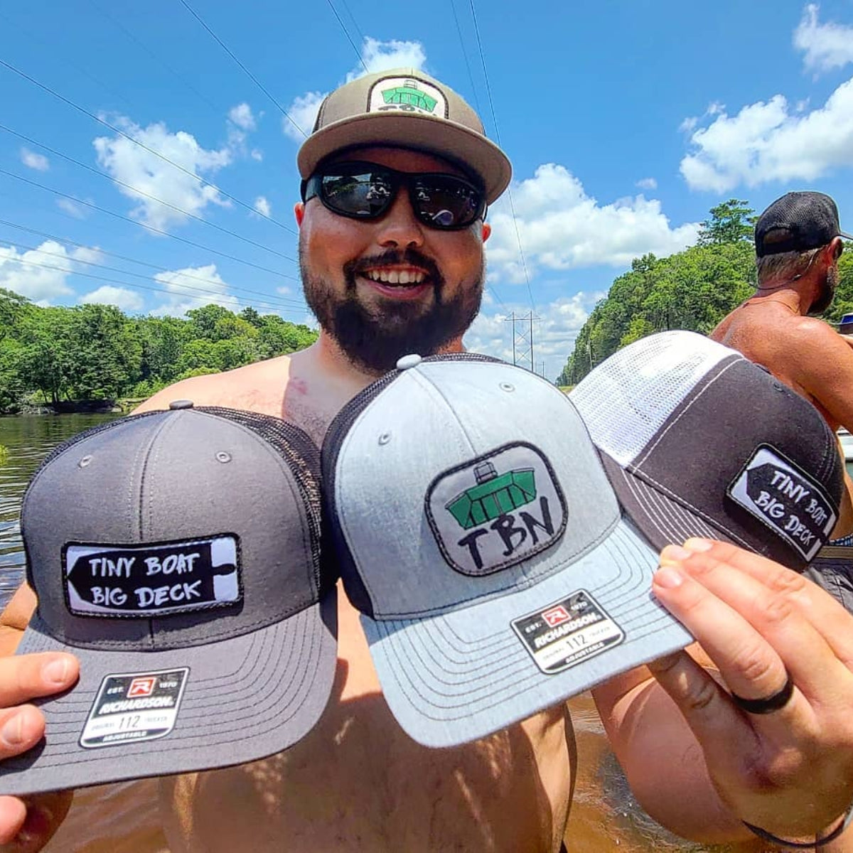 Man holding three Richardson 112 hats: charcoal with "Tiny Boat Big Deck" patch, light gray with "TBN Jon Boat" patch, and black/white mesh with "Tiny Boat Big Deck" patch.