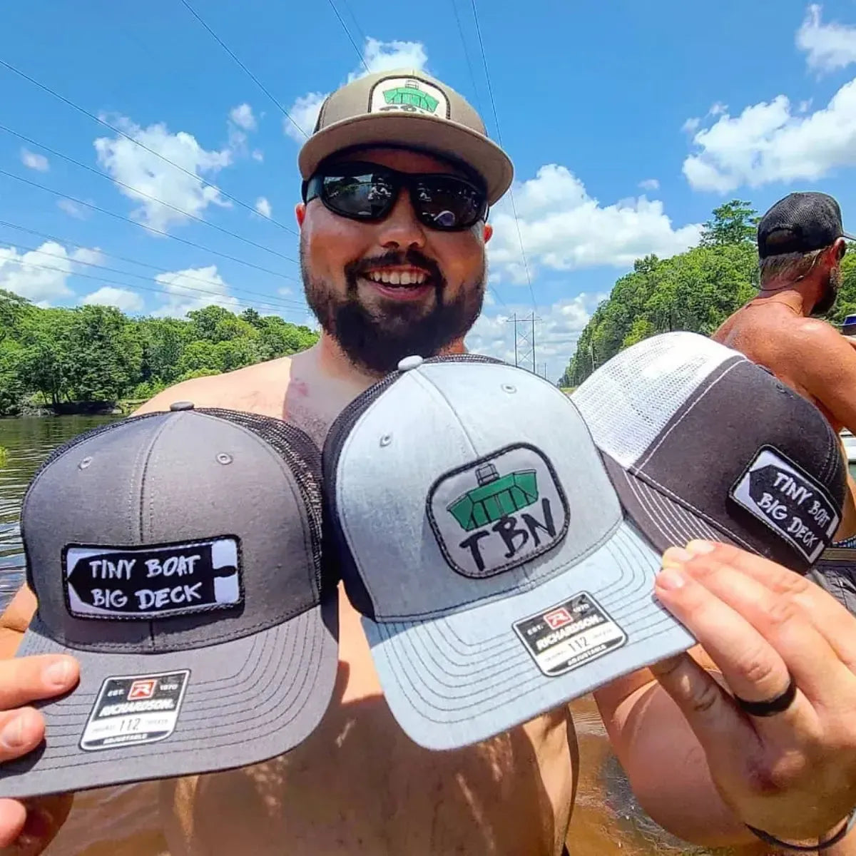 Man holding three Richardson 112 hats: charcoal with "Tiny Boat Big Deck" patch, light gray with "TBN Jon Boat" patch, and black/white mesh with "Tiny Boat Big Deck" patch.