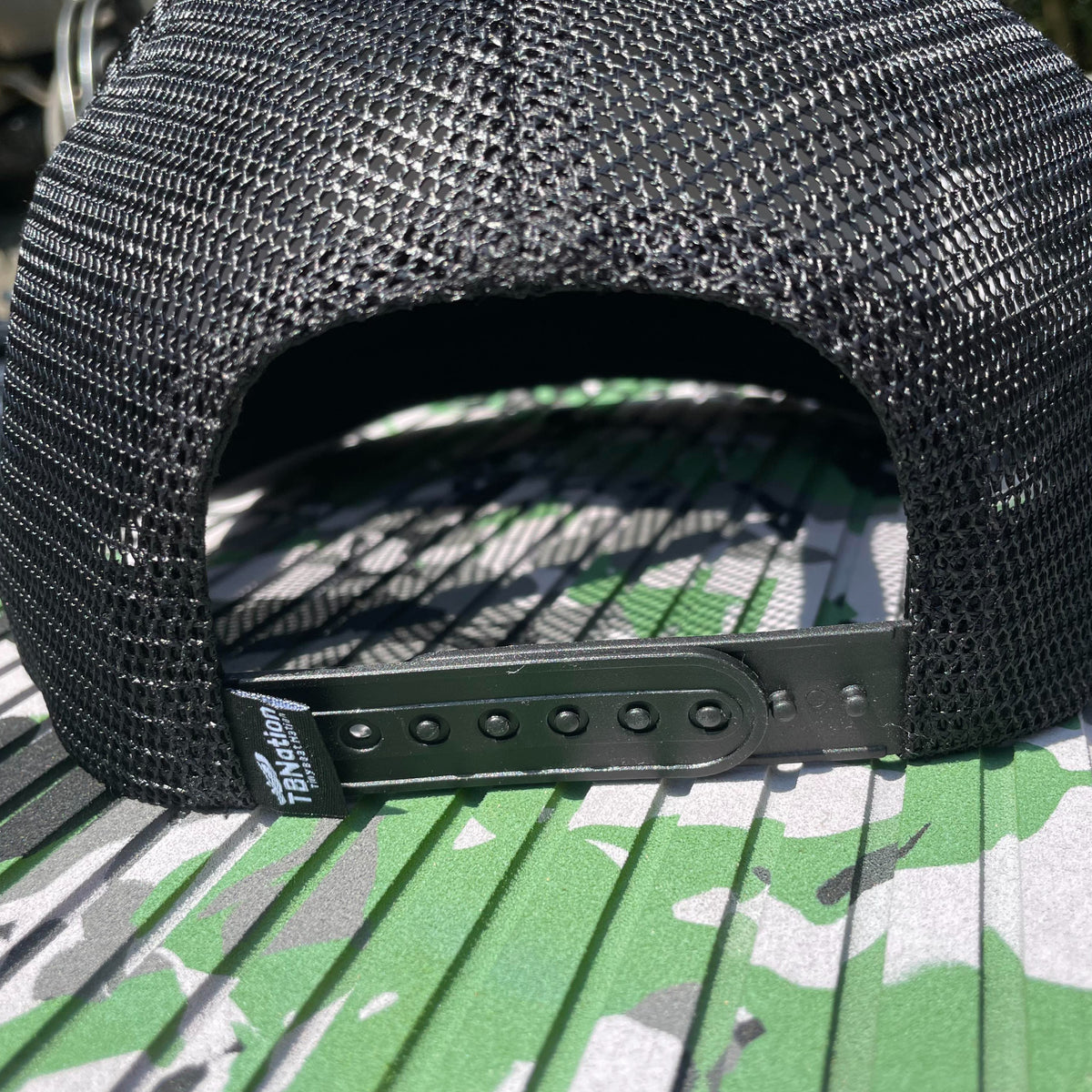 Black mesh trucker hat with snapback closure and a "TBNation" tag, resting on a green and grey patterned surface.