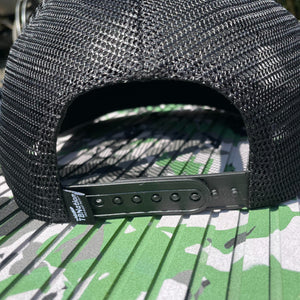 Black mesh trucker hat with snapback closure and a "TBNation" tag, resting on a green and grey patterned surface.