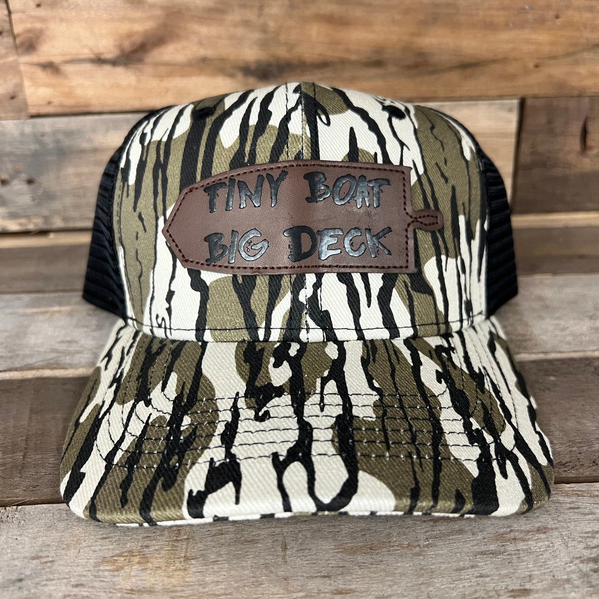 Camo trucker hat with black mesh back and a brown leather patch reading "Tiny Boat Big Deck.