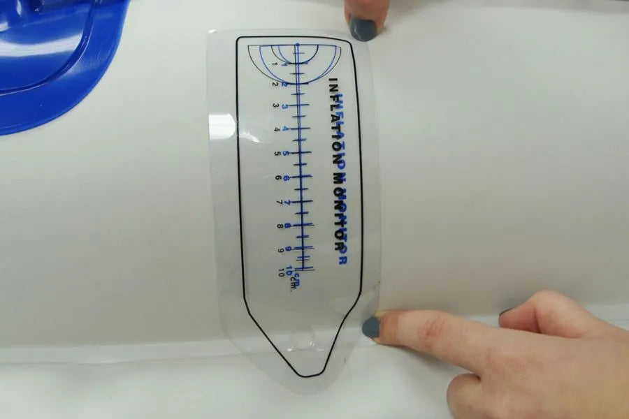 Clear pliable plastic inflation gauge with blue markings, held against a white kayak surface.