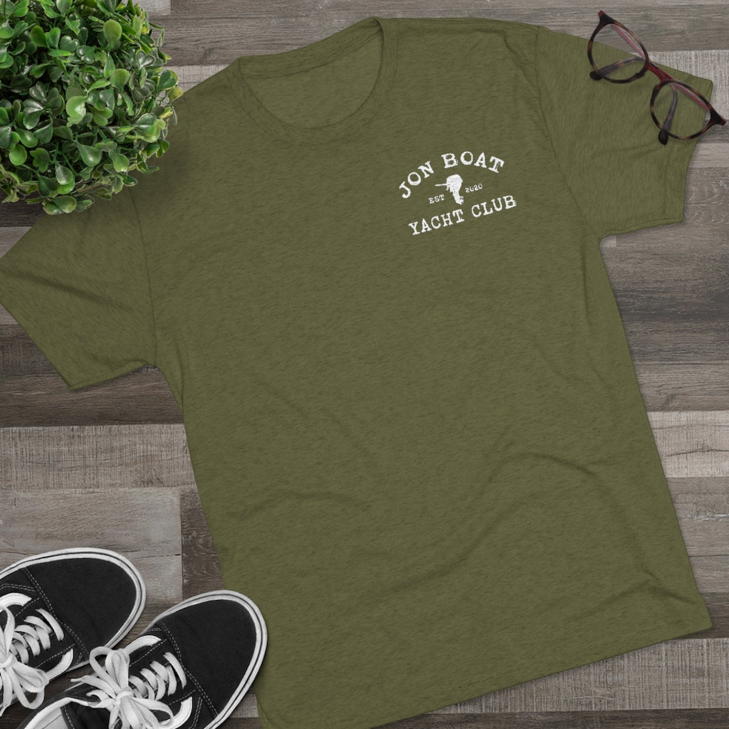 Olive green tri-blend tee with "Jon Boat Yacht Club" logo, black sneakers, plant, and glasses on wood.