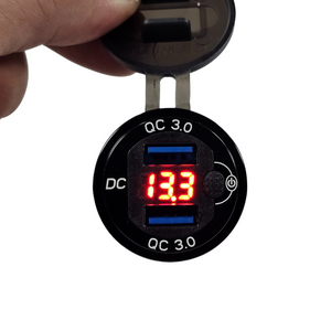 Black LED voltmeter with red digital display and dual blue QC 3.0 USB ports, held by a hand.