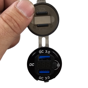Black round LED voltmeter with dual blue QC 3.0 USB ports, held by fingers.