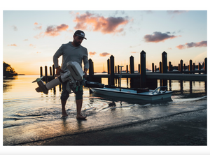 Man carries a gray Newport NT600 6hp electric outboard motor into the water at sunset, next to a small boat.