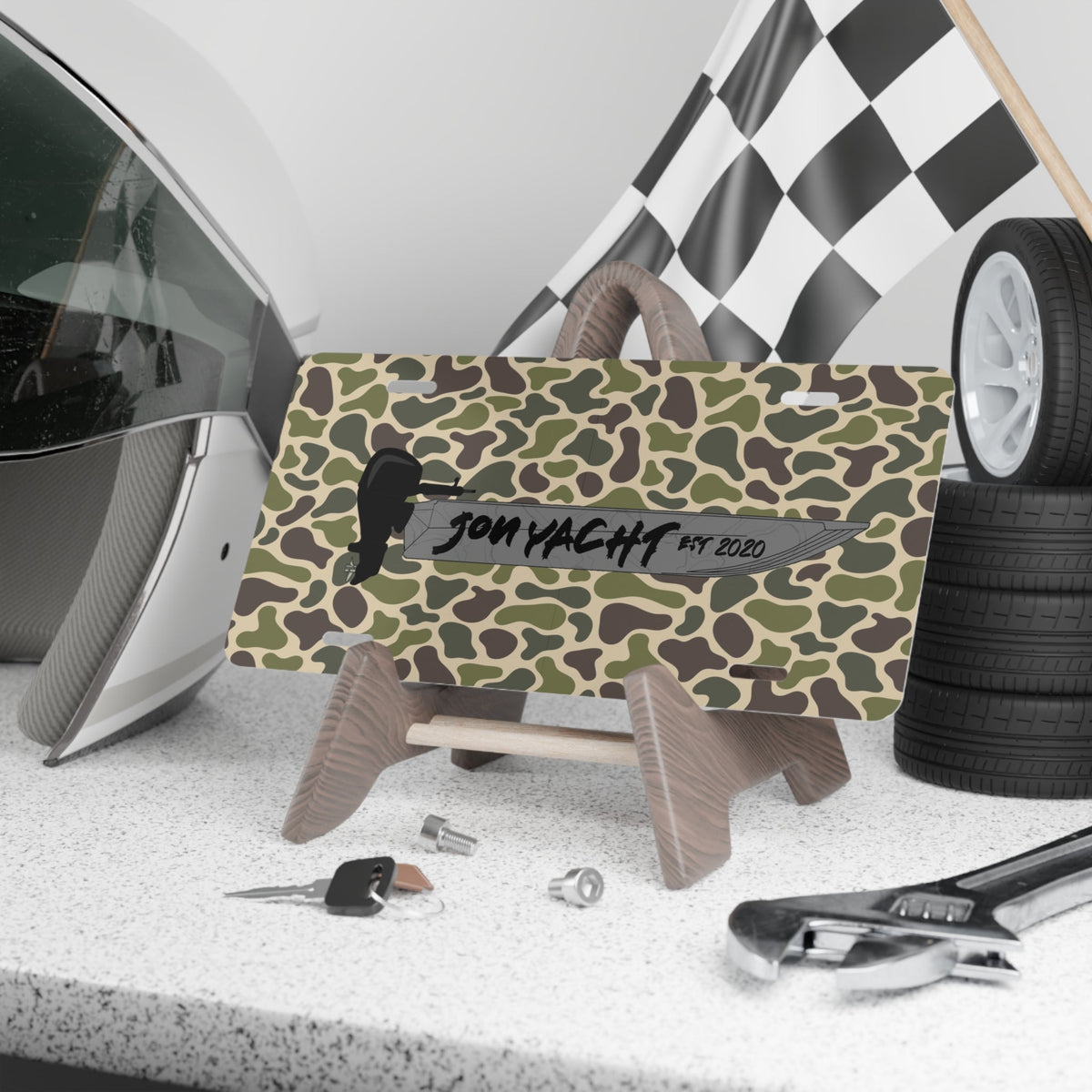 Camo aluminum license plate with "JON YACHT EST 2020" text and boat graphic, displayed on a stand.