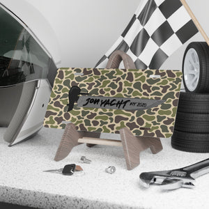 Camo aluminum license plate with "JON YACHT EST 2020" text and boat graphic, displayed on a stand.
