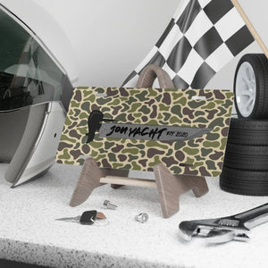 Camo aluminum license plate with "JON YACHT EST 2020" text and boat graphic, displayed on a stand.
