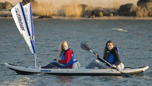 Two women in a gray Sea Eagle FastTrack inflatable kayak with a white and blue sail, paddling on calm water.