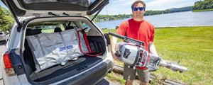 Man holding a silver outboard motor next to a white SUV with a packed Sea Eagle inflatable boat.