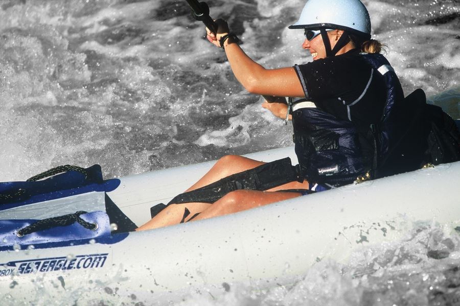 Smiling kayaker in a white helmet and black shirt, using black Sea Eagle thigh straps in a white inflatable kayak on choppy water.