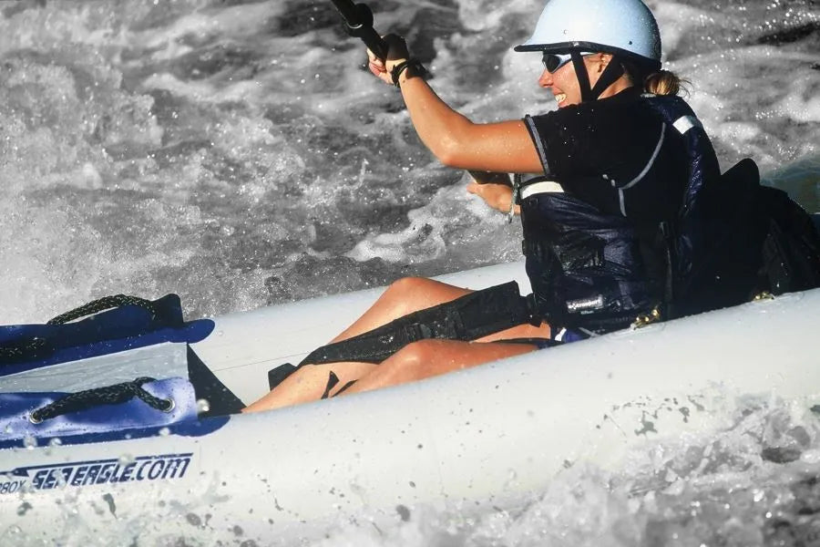 Smiling kayaker in a white helmet and black shirt, using black Sea Eagle thigh straps in a white inflatable kayak on choppy water.