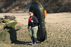 Man carrying a rolled-up black and green inflatable kayak on his back with orange straps and a paddle.